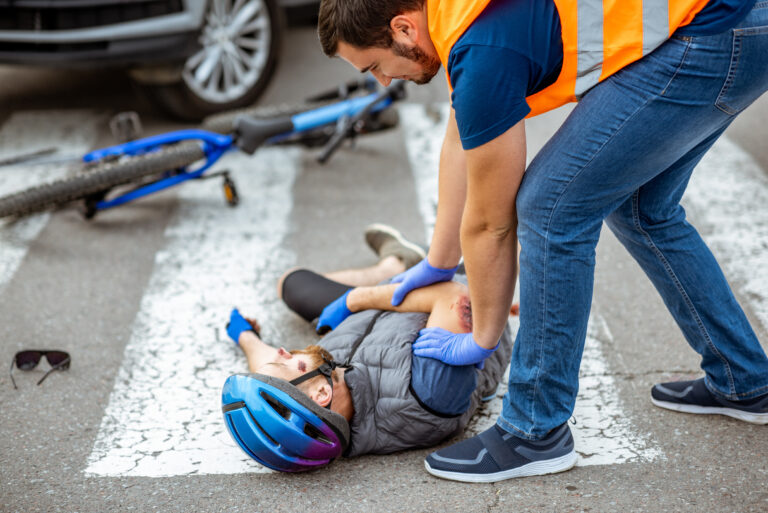 Road accident with injured cyclist lying on the pedestrian crossing near the bicycle and car, male driver providing first aid